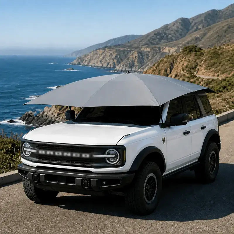 Automatic car umbrella silver gray deployed on white Ford Bronco SUV on scenic coastal highway with ocean and mountain backdrop